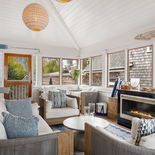 A cozy sunroom with white wooden walls, a fireplace, seating with blue cushions, decorative pieces, and large windows with an outdoor view.