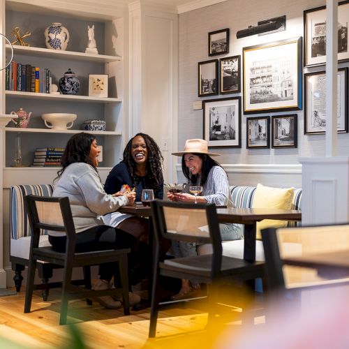 Three people are joyfully chatting at a cozy table in a warmly decorated room with shelves and framed pictures on the wall.