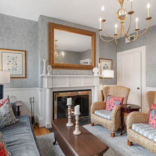 A cozy living room with a fireplace, wicker chairs, a gray couch, colorful pillows, elegant chandelier, and decorative art pieces on walls.