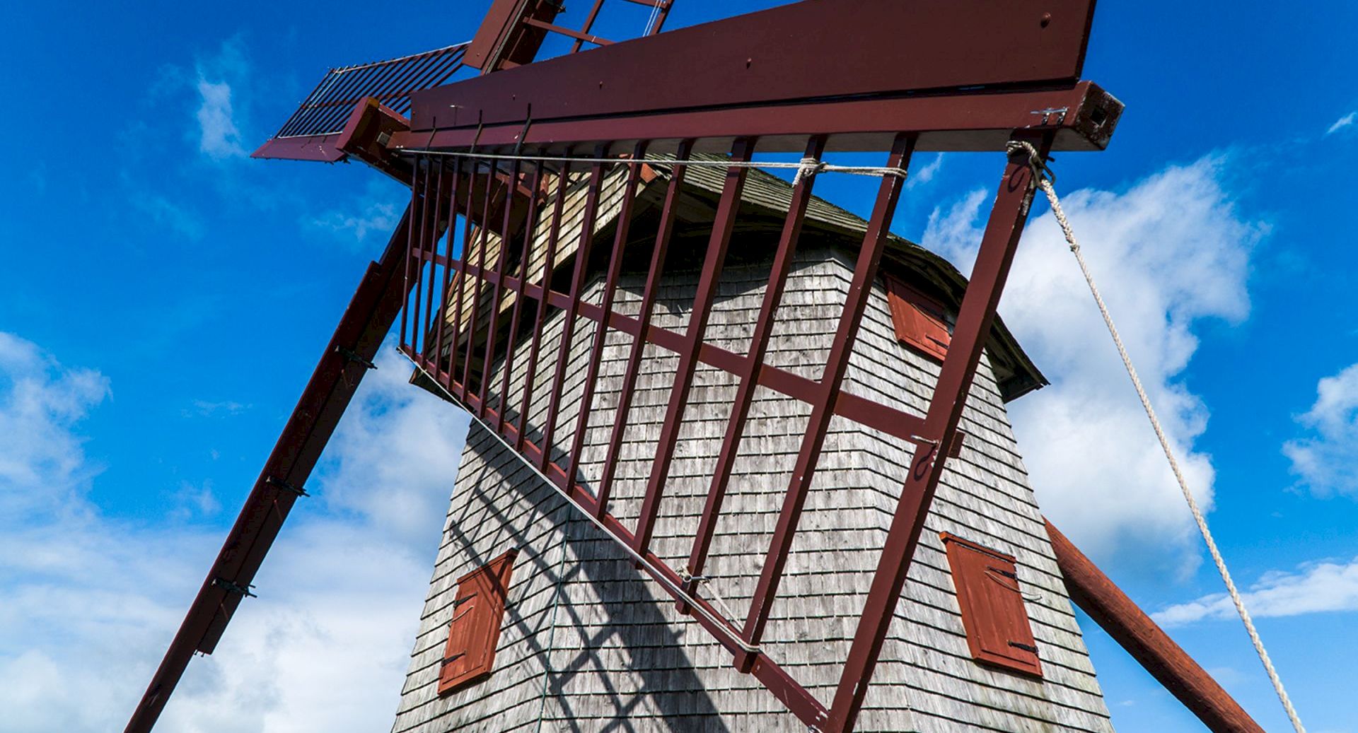 A tall wooden windmill with red blades and chains against a bright blue sky with scattered clouds.