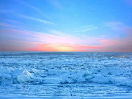 A frozen ocean with snow-covered ice fields stretches to the horizon under a colorful dawn sky.