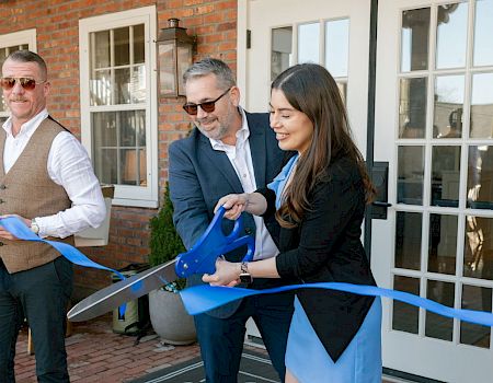 People in suits cut a blue ribbon outside a brick-and-white building, celebrating an opening with smiles.