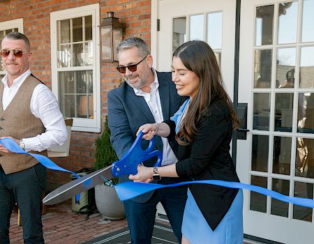 Three people are cutting a blue ribbon with large scissors at an event in front of a building.