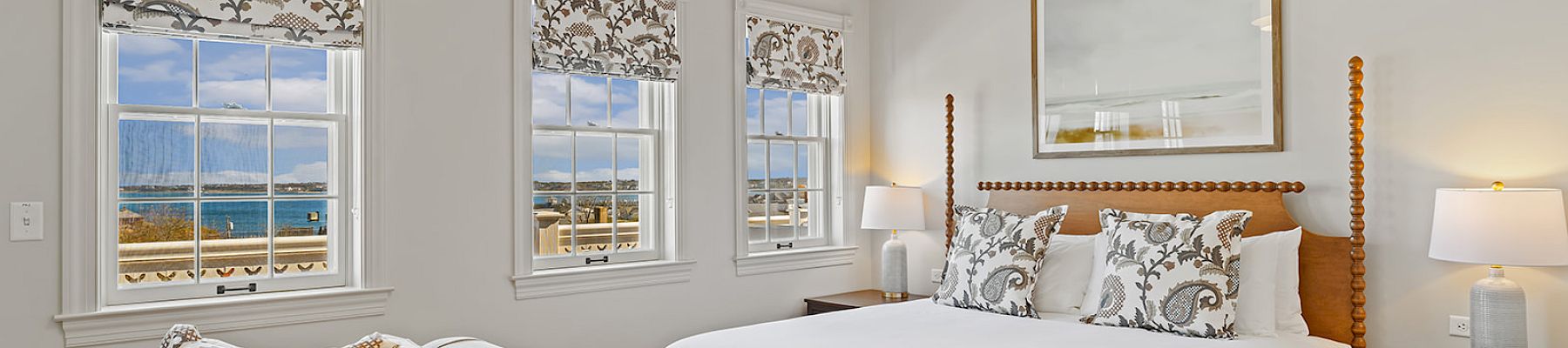A cozy bedroom features a large bed, decorative pillows, patterned blinds, and a scenic view of the ocean through the windows.