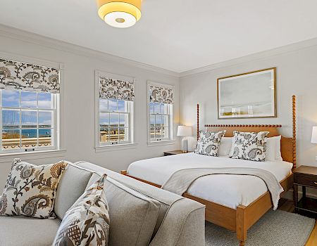 A cozy bedroom features a large bed, decorative pillows, patterned blinds, and a scenic view of the ocean through the windows.