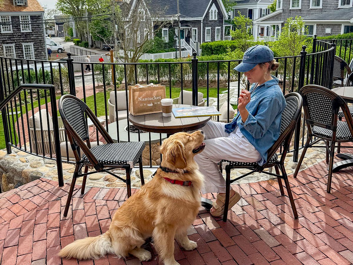 A woman on a patio enjoys a drink with her golden retriever while quaint houses sit across the street.