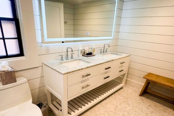 Modern bathroom with double sink vanity, large mirror, toilet, tissue box, and wooden bench, all set against light wood paneling.