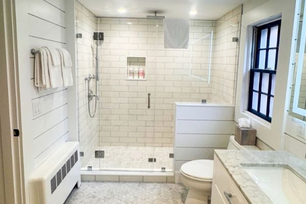 A modern bathroom with a walk-in shower, white tiles, vanity with a sink, mirror, and toilet, featuring sleek and clean design elements.