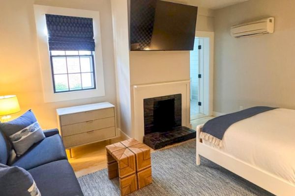 A cozy bedroom features a bed, sofa, wall-mounted TV, and fireplace, with a dresser and lamp under a window for a modern touch.