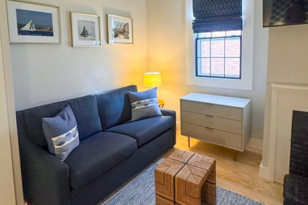 A cozy living room with a dark sofa, patterned pillows, a small wooden table, framed photos, window with blinds, and a dresser.