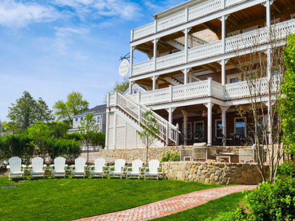 Large building with multiple wooden balconies, green lawn, white chairs, and a stone pathway under a blue sky.