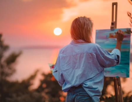 A person is painting on a canvas during a sunset, with a vibrant sky and blurred natural landscape in the background.