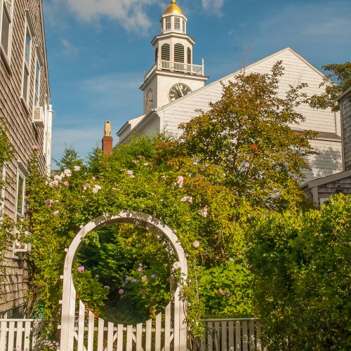 A charming garden path with a white picket fence leads to a building with a clock tower under a blue sky, surrounded by lush greenery.