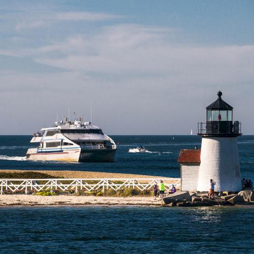 A lighthouse stands on a sandy point with a white fence, as a large boat cruises in the background on a sunny day.