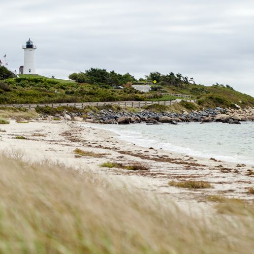 A lighthouse and house on a grassy hill overlook a rocky beach with calm waves under a cloudy sky.