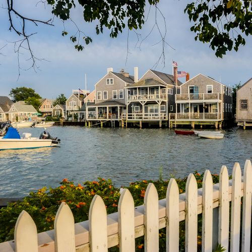 A waterfront scene with houses, boats on the water, and a white picket fence in the foreground under a tree.
