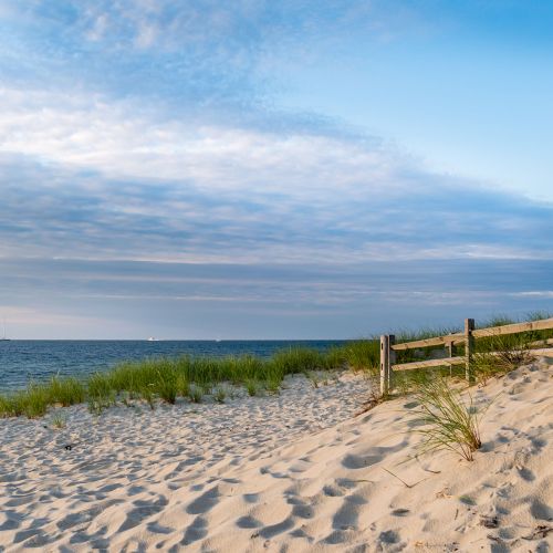 Sunny sandy beach with dunes, grasses, a wooden rail, calm blue ocean, and a cloudy sky stretching to the horizon.