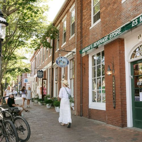 A sunny storefront street with brick buildings and signs like &ldquo;Murray&rsquo;s Beverage Store,&rdquo; people walking, and a bicycle parked by a tree.