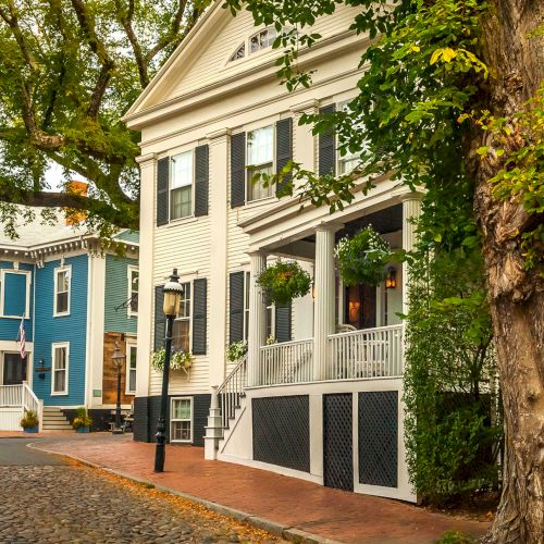 A quiet street lined with historic houses, white and blue facades, porches, shutters, and leafy trees casting shade.