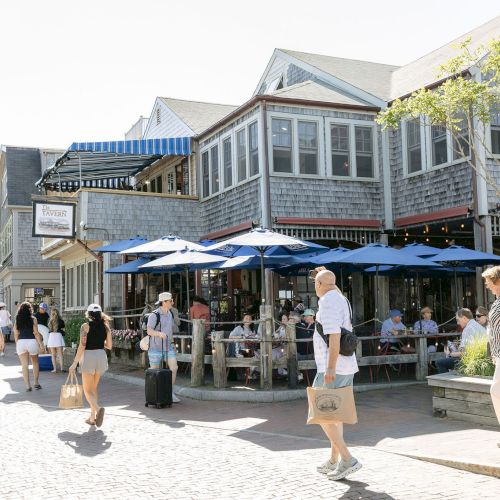 An outdoor restaurant patio with blue umbrellas and people gathered outside a rustic tavern building on a sunny street.