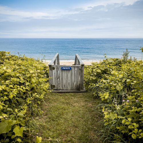 A private wooden gate stands among green shrubs, opening to a calm ocean and cloudy sky on the coast.