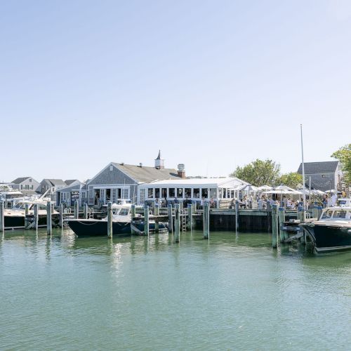 Boats are docked at a sunny marina with people gathered near white tented event areas along the waterfront.