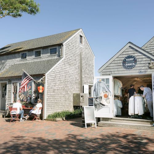 Outdoor storefronts at a seaside shop, with people browsing T-shirts and flags under a sunny sky.