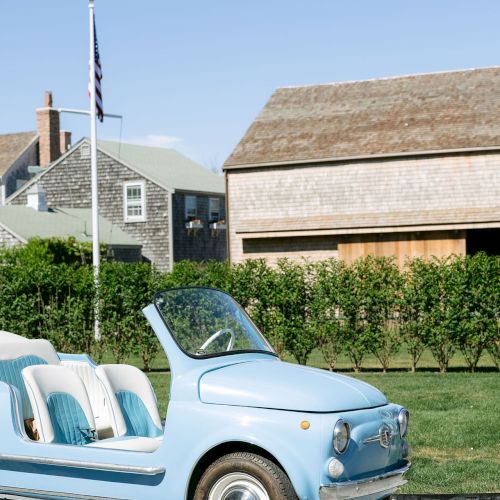 A light-blue vintage convertible sits outdoors beside manicured hedges and a flagpole in front of coastal-style homes.