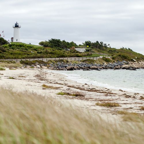 A white lighthouse stands on a rocky, grassy coast above sandy dunes, with waves and a cloudy sky rolling in beside it.
