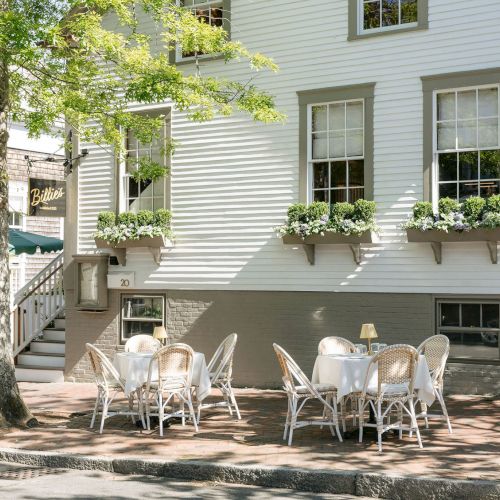 An outdoor patio with tables, chairs, planters, and a white building with windows plus a &ldquo;Billie&rsquo;s&rdquo; banner.