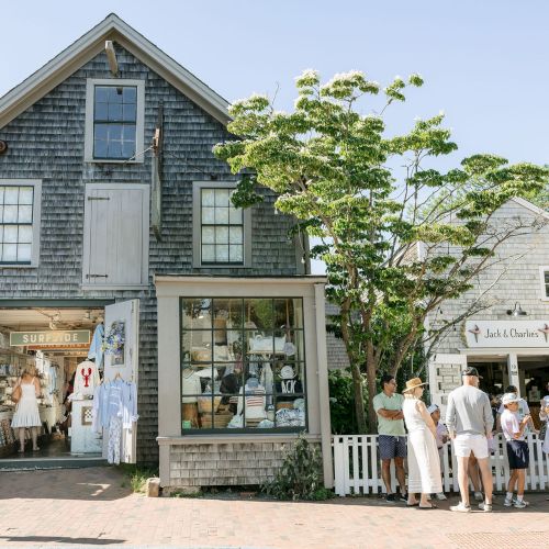 A seaside shopping street with storefronts, people browsing outside &ldquo;Jack & Charli&eacute;s&rdquo; and &ldquo;Surfside,&rdquo; under sunny skies.
