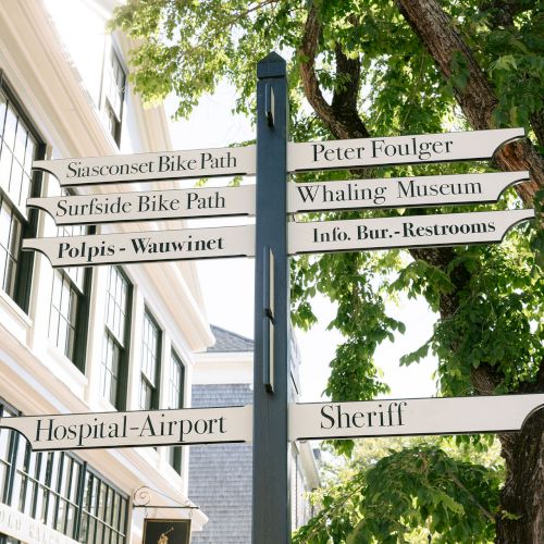 A street signpost lists routes like Siasconset Bike Path, Whaling Museum, restrooms, Hospital-Airport, and Sheriff beneath trees.
