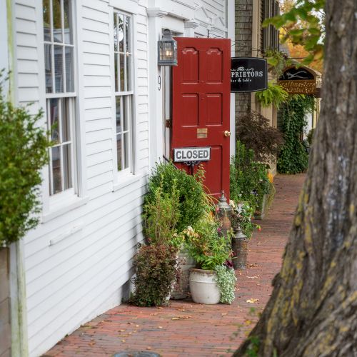 A red door marked "closed," surrounded by potted plants, is set against a brick path and white building exterior with signs.