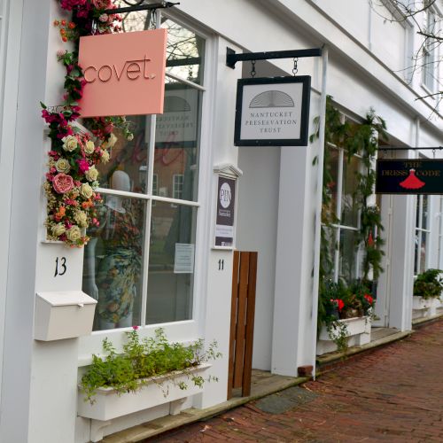 A row of shops with signs, floral decorations, and large windows lines a brick-paved street.