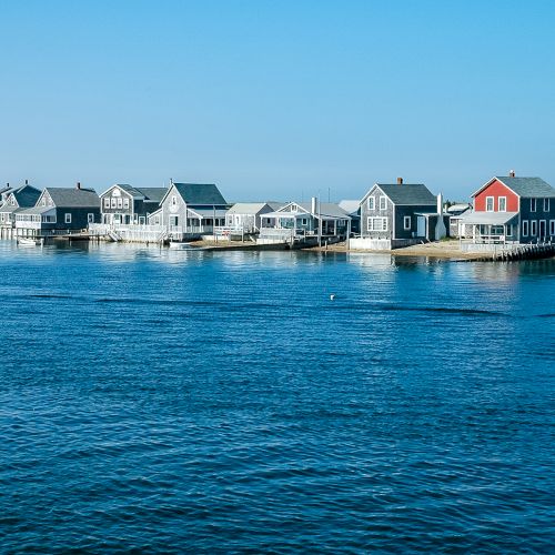 A row of coastal houses, with one red, lines a waterfront on a sunny day under a clear blue sky.