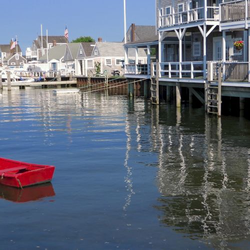 A red boat floats near waterfront buildings with decks over the water, casting reflections in a calm, clear harbor setting.