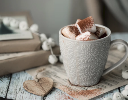 A decorative mug filled with hot chocolate and marshmallows on a table, surrounded by books, a wooden heart, and a string of decorations.