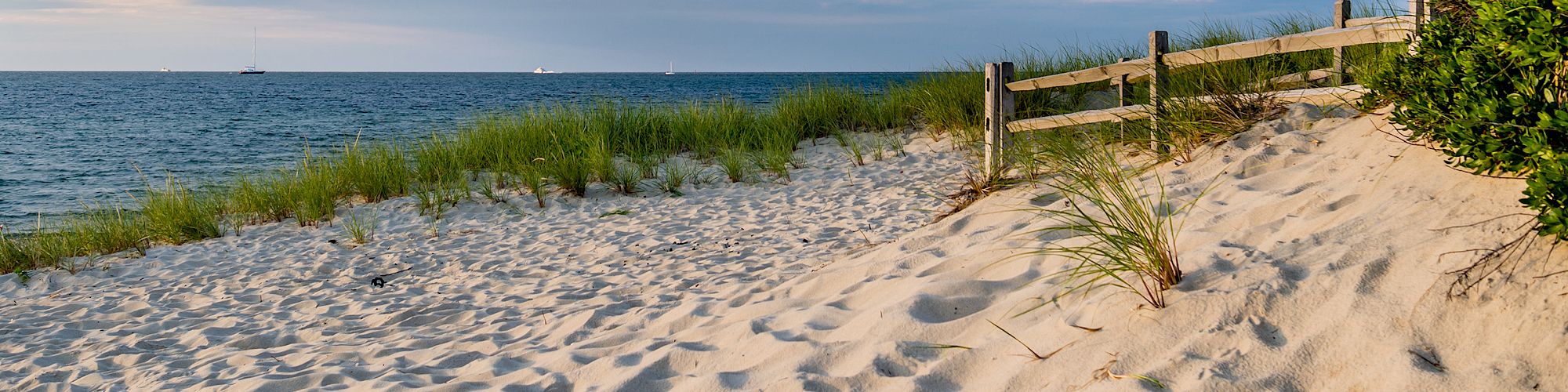 A sandy beach with light dunes, a wooden fence, sparse grasses, calm sea, and a blue sky with gentle clouds, peaceful coastal scene.
