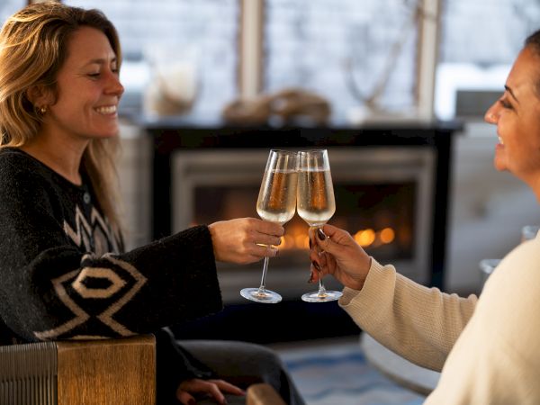 Two women share a cozy moment, clinking wine glasses near a fireplace as they chat and smile warmly.