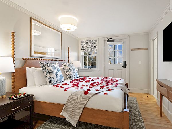 A cozy hotel-style bedroom with a wooden bed, white linens, blue patterned pillows, rose petals on the bed, a wall-mounted TV, and a dresser.