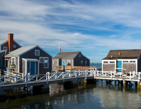 Colorful ocean-side cottages on stilts connected by a white walkway, with flags, blue skies, and calm water beneath.