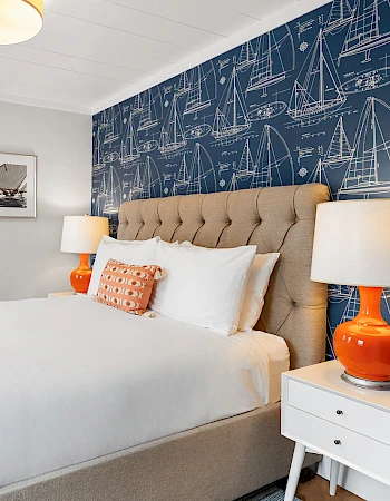 A bright hotel room with a tufted beige headboard, blue sailboat wallpaper, white bedding with orange accents, two white nightstands, orange lamps, and a small sitting area near a window.