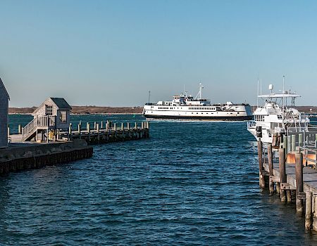 A harbor scene with calm blue water, a pier and small gray buildings on the left, and a large white ferry docked in the distance, under a clear sky.