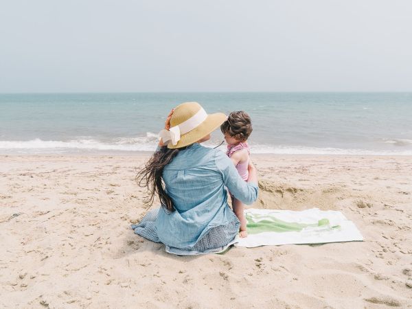 A woman in a floppy hat sits on a beach blanket with a child, watching the sea as gentle waves roll in on a sunny day.