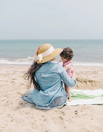 A woman wearing a sun hat and denim jacket sits on a beach blanket with a child, gazing at the calm sea under a clear sky.