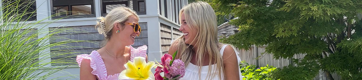 Two women in summer dresses stroll a garden path, holding colorful bouquets and chatting happily near a bright porch.