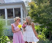 Two women in summer dresses stroll a garden path, holding colorful bouquets and chatting happily near a bright porch.