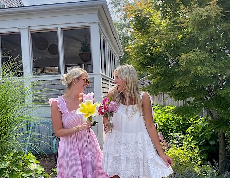 Two women in summer dresses stroll a garden path, holding colorful bouquets and chatting happily near a bright porch.