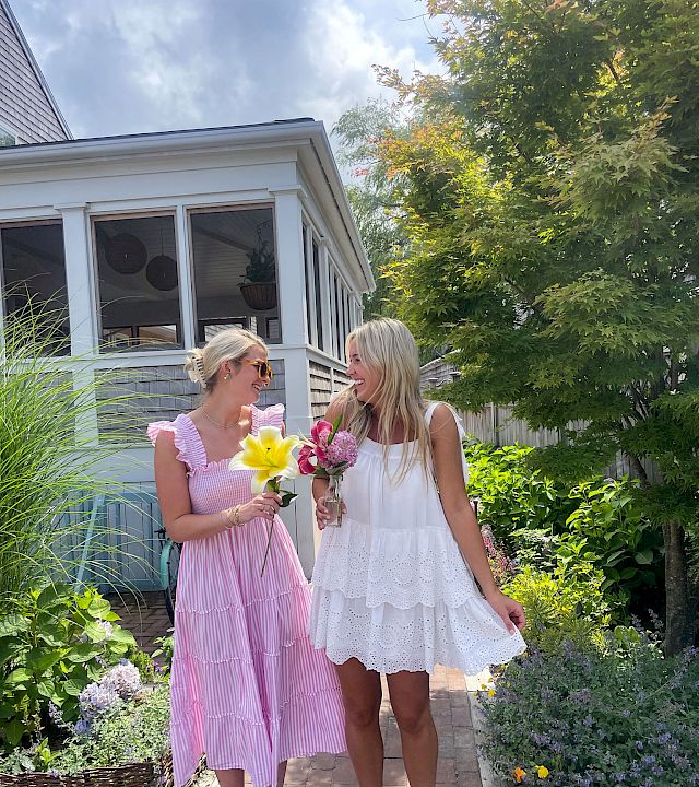 Two women in summer dresses stroll a garden path, holding colorful bouquets and chatting happily near a bright porch.