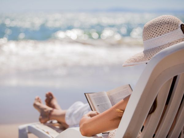 A person in a sun hat relaxing on a beach chair by the ocean, reading a book as waves roll in.